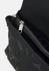 Schwarze Handtasche mit einer strukturierten Außenseite und floralen Prägungen. Das Innere umfasst ein mit Reißverschluss versehenes Fach und ein schwarzes Futter. Silberne Reißverschlussbeschläge.