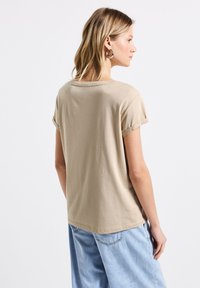 Camiseta de algodón beige con mangas cortas enrolladas, un escote redondo y un corte relajado, mostrada desde una vista lateral y posterior.