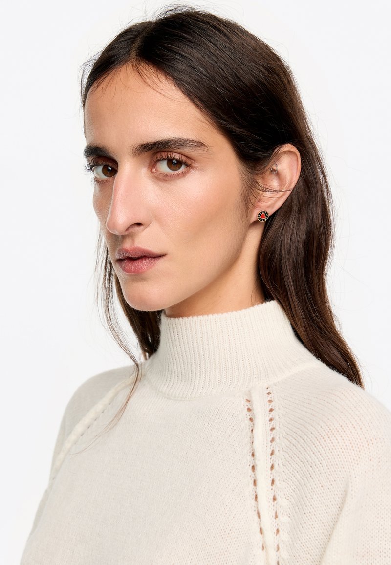 Pendientes con un diseño circular que presenta un fondo negro y un motivo floral en rojo. El modelo lleva un suéter de cuello alto de color crema con textura en canalé.