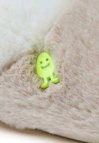 Un pelage beige doux avec un petit personnage en plastique vert vif, souriant et incrusté dedans, ajoutant un détail ludique à la texture.