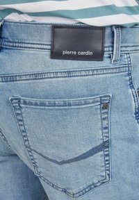 Helle blaue Denim-Jeans mit einem Lederetikett, das "pierre cardin" auf dem Bund trägt, und einer geschwungenen Rückentaschen-Design.