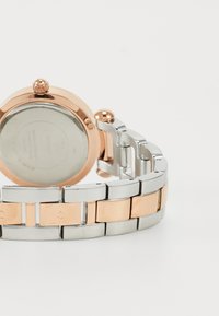 Montre en argent et en rose gold avec un bracelet en métal. Boîtier rond avec un fond transparent et une couronne décorative. Les maillons varient en couleur et en finition.