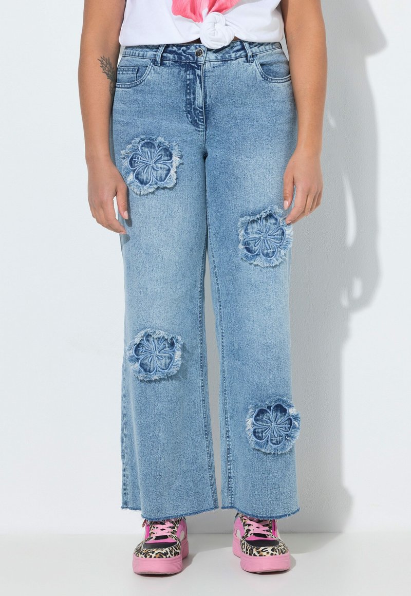 Vaaleansiniset leveälahkeiset farkut, joissa on kuluneet kukkakuviolliset paikat, yhdistettynä vaaleanpunaisiin leopardikuvioisiin korokepohjakenkiin ja solmittuun valkoiseen t-paitaan.