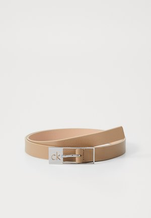 Világos bézs bőr öv sima textúrával. Ezüst fém csattal rendelkezik, amelyen a "ck" felirat ki van préselve, és minimalista dizájn jellemzi.