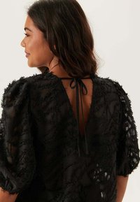Blusa nera con texture e maniche a palloncino, caratterizzata da una profonda scollatura a V sul retro e un fiocco decorativo. Realizzata in tessuto leggero e trasparente con motivi cuciti.