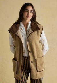Beige Fleece-Weste mit braunem Cordkragen, ausgestattet mit zwei Fronttaschen, offenem Design und umrandeten Kanten. Gestreifte Hose darunter.