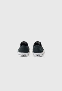 Sorte semska skoene med lavt design, hvite gummisåler og "CONVERSE ALL★STAR" merkevaren på hælen. Ingen synlige mønstre.