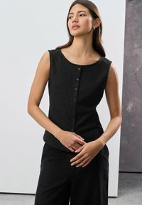 Black sleeveless top features a textured fabric, scoop neckline, and front button closure, paired with high-waisted black pants.