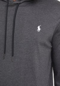 Grijze hoodie met een trekkoord capuchon. Bevat een klein wit logo op de borst. De stof lijkt zacht met een subtiele textuur.