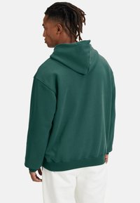Felpa verde con cappuccio realizzata in tessuto morbido. Presenta una vestibilità comoda e polsini a coste, con una finitura testurizzata e tasca a marsupio sul davanti.