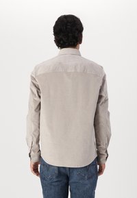 Camisa de pana beige de manga larga, con cuello abotonado y canalones verticales. Presenta un dobladillo redondeado y puños abotonados.