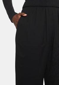 Pantaloni neri in tessuto con vita elasticizzata, tasche laterali e una texture liscia, dal taglio comodo e morbido. Mano posizionata in modo casuale nella tasca.