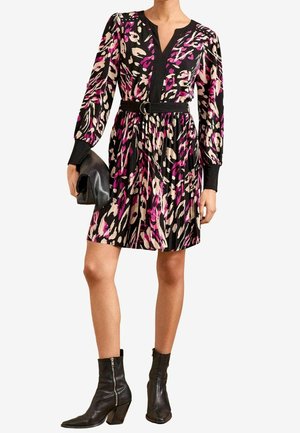 Kvinde iført en kort sort kjole med pink og beige abstrakt blomsterprint, sort bælte, holder en sort clutch og bærer sorte ankelstøvler.