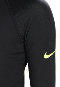 T-shirt de sport noir à manches longues avec une coupe ajustée, arborant un logo Nike jaune sur la manche gauche et un tissu lisse et texturé.