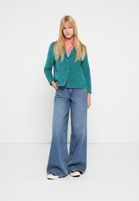 Turkusowa marynarka ze sztruksu z przednimi kieszeniami noszona na czerwono-białej koszuli w kratę, w połączeniu z szerokimi niebieskimi jeansami i czarnymi tenisówkami.