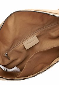 Interior de bolsa de tela beige que muestra un bolsillo con cremallera con una etiqueta con el logo de Tamaris y un bolsillo abierto, todo dentro de un compartimento principal sin cerrar.
