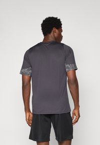 T-shirt de sport gris à manches courtes avec une bordure à motifs sur les poignets, associé à un short noir. Texture lisse, design ajusté.