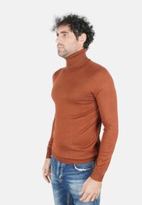 Uomo con capelli ricci che indossa un dolcevita color ruggine e jeans blu, in piedi davanti a uno sfondo bianco semplice.