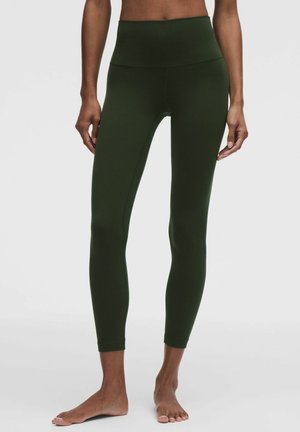 Persoon draagt een donker groene legging met hoge taille en volledige lengte, staat blootsvoets tegen een effen lichte achtergrond.