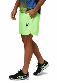 Pantaloni sportivi di un brillante verde neon con tasche e una fessura laterale, abbinati a scarpe da corsa nere con accenti verdi.