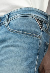 Ανοιχτό μπλε τζιν παντελόνι με ελαφρώς φθαρμένη υφή. Διαθέτει μια μαύρη ετικέτα με λευκό κείμενο και μια ροζ λεπτομέρεια στην πίσω τσέπη.