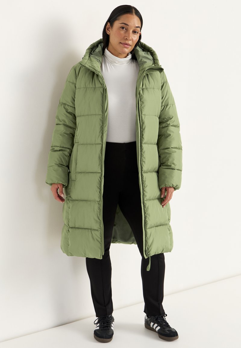 Kvinde iført en lang grøn pufferjakke med hætte, hvid turtleneck, sorte bukser og sorte sneakers med hvide striber, stående mod en hvid væg.