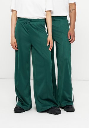 Deux personnes portant un pantalon large vert foncé avec des bandes blanches latérales et des chaussures noires épaisses, debout proches l'une de l'autre.