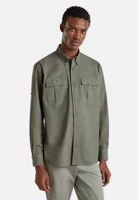 Camisa de botones verde oliva con cuello, dos bolsillos frontales y mangas largas. Textura suave y corte ajustado.