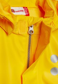 Gul vattentät jacka med hög krage, YKK-dragkedja, kardborrestängning och silverfärgade cirkulära detaljer framtill.