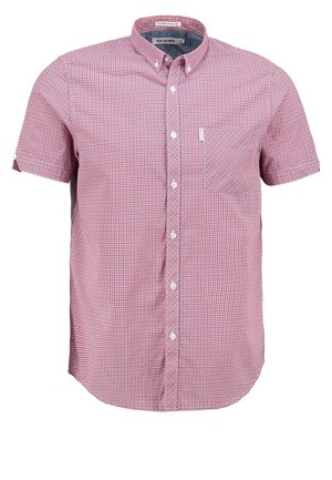 Mann trägt kurzärmeliges rotes kariertes Hemd, schwarze Hose, blaue Slip-On-Schuhe und trägt eine braune Leder-Umhängetasche mit Fronttaschen und Reißverschlüssen.