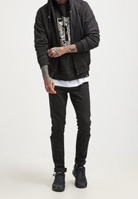 Uomo che indossa una felpa nera con zip, una t-shirt nera con grafica, jeans skinny neri strappati e sneakers nere, con tatuaggi su braccia e mani.