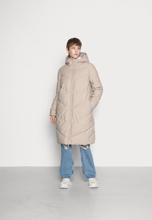 Doudoune beige clair avec capuche, dotée d'une fermeture éclair et d'un design matelassé en chevrons. Associée à un jean bleu déchiré et des baskets.