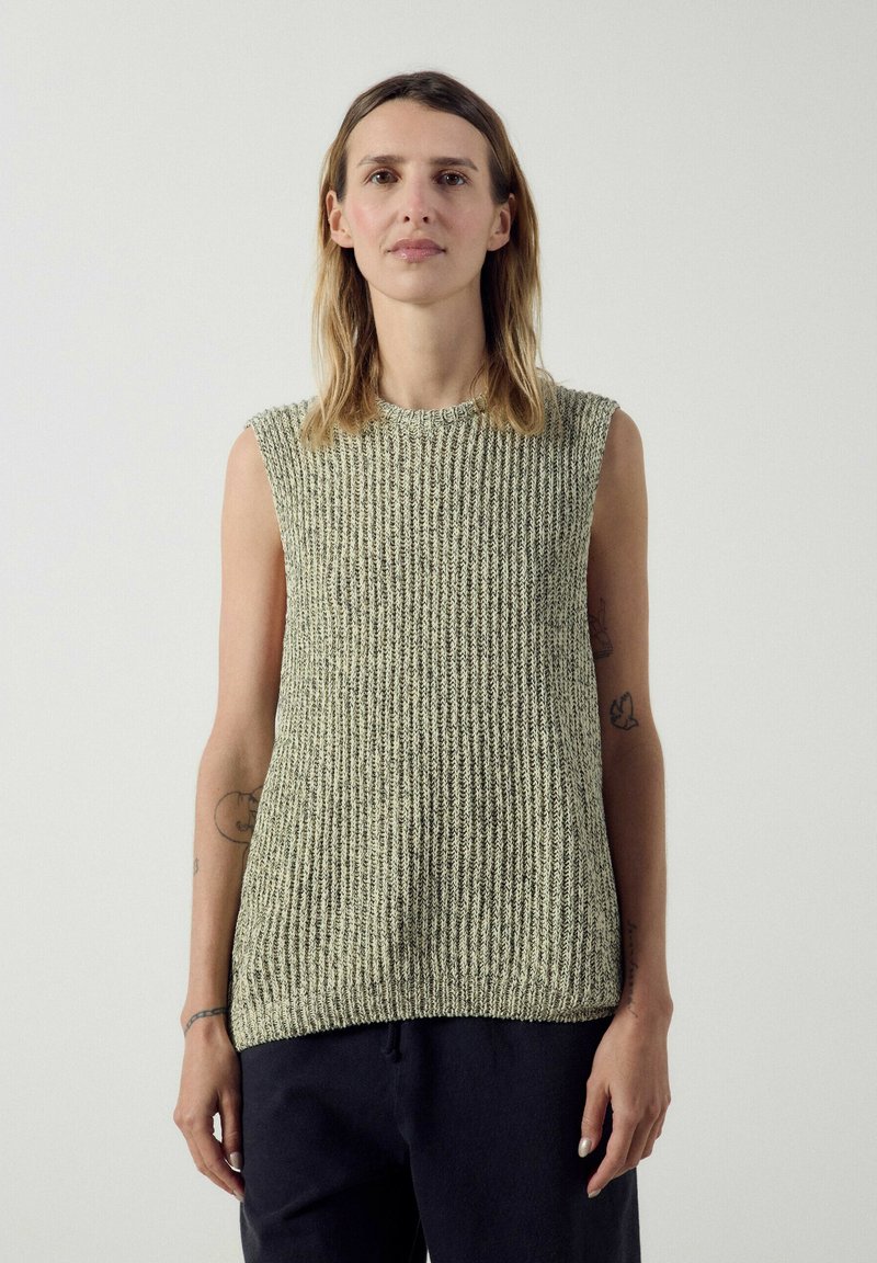 Femme aux cheveux mi-longs portant un haut en tricot texturé sans manches vert clair et un pantalon foncé, debout devant un fond clair uni.