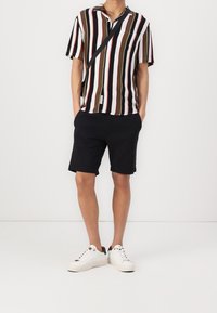 Korte mouwen overhemd met verticale strepen in olijf, zwart, rood en wit, gecombineerd met zwarte shorts en witte sneakers.