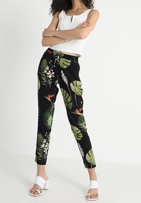 Zwarte bloemenbroek met groene bladeren en witte bloemen, met een elastische tailleband en een enkel-lange pasvorm. Gecombineerd met witte block-heeled sandalen.