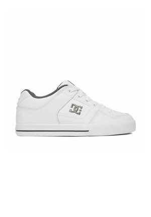 Zapatilla blanca de corte bajo con forro negro, cierre con cordones y logo DC negro en el lateral sobre una suela blanca con una delgada franja negra.