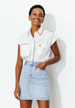 Femme souriante portant une chemise blanche à manches courtes avec une broderie colorée et une jupe en denim bleu clair, debout devant un fond blanc.