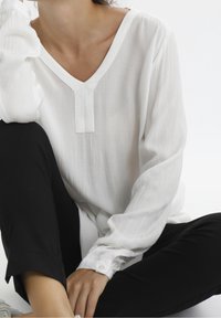 Blouse blanche à manches longues avec un col en V et un tissu texturé, dotée d'un détail de bouton sur le poignet, associée à un pantalon noir.