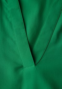 Tessuto verde con scollatura a V e texture liscia. Cuciture visibili che accentuano i bordi e le cuciture.