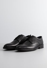Chaussures de ville en cuir noir avec une finition lisse, présentant un panneau en maille texturée et un design classique à lacets ; bout arrondi et semelle plate.
