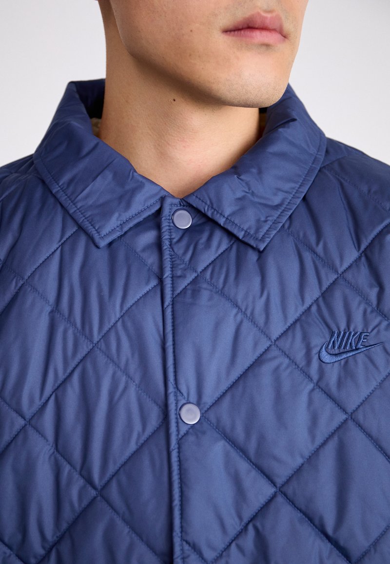 Veste matelassée navy avec col en pointe, boutons-pression et logo Nike brodé dans le même fil. Texture lisse avec motif en losanges.