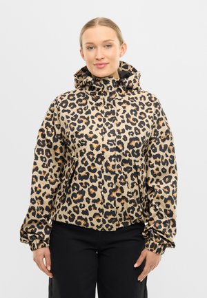 Femeie purtând o jachetă cu glugă bej cu imprimeu de leopard negru și manșete elastice, plus pantaloni negri, stând pe un fundal simplu.