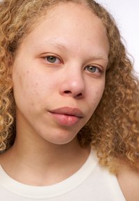 Close-up van een persoon met krullend, lichtbruin haar, een lichte huid en lichte sproeten, in een witte tanktop. Natuurlijke make-up met zachte lippen.