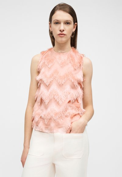 Γυναίκα με ίσια καστανά μαλλιά που φορά χρυσούς κρίκους στα αυτιά, ένα αμάνικο ανοιχτό ροζ τοπ με υφή και λευκό παντελόνι, με το ένα χέρι στην τσέπη.