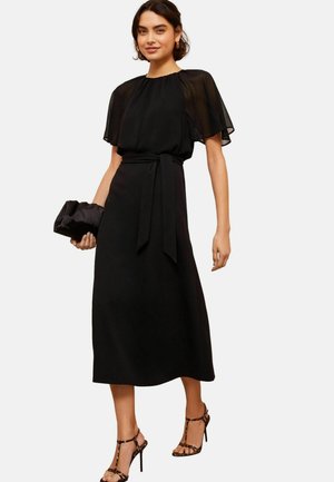 Abito lungo nero con maniche corte trasparenti, scollo arricciato e cintura in vita. Abbinato a sandali con cinturino e una borsa a tracolla nera.
