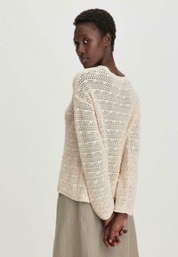 Hellbeige gestrickter Pullover mit einem offenen Maschenmuster, Rundhalsausschnitt und langen Ärmeln, der vor einem hellen Hintergrund liegt.