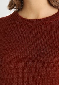 Długi, rustykalny sweter w kolorze rdzy, wykonany na drutach z prążkowaną fakturą i okrągłym dekoltem, prezentujący miękką, przytulną tkaninę.