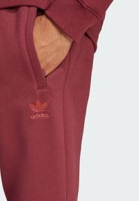 Bordové tepláky majú bočnú vrecko a výšivku oranžového loga Adidas na stehne. Tkanina má hladký, textúrovaný povrch.