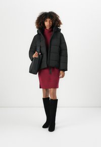 Geacă puffer neagră cu glugă, rochie-sweater texturată de culoare bordo cu motive în formă de diamant și cizme negre până la genunchi. Modelul ține o geantă de mână neagră.