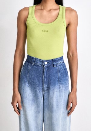 Woman wearing a lime green ribbed tank top with "BOSS" logo and high-waisted blue ombre denim jeans against a plain white background.